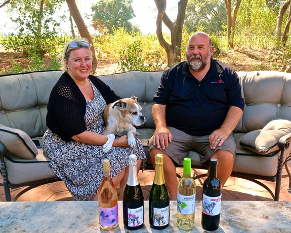 man and woman sitting on a chair with dog on womans lap and wine on the table
